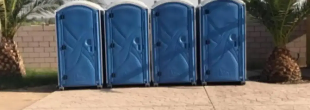 Four blue standard porta potties lined up on a paved lot with palm trees behind them